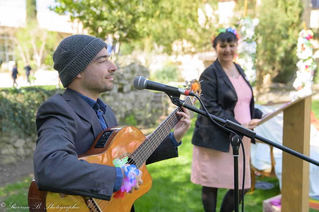 Le chanteur Nicolas Demailly (nicolasdemailly.fr) en premier plan avec sa guitare, et l’officiante Frédérique de Cœurs au diapason en arrière‑plan flou, lors du Festival des 5 sens au Mas de la Baraque de Sérignac