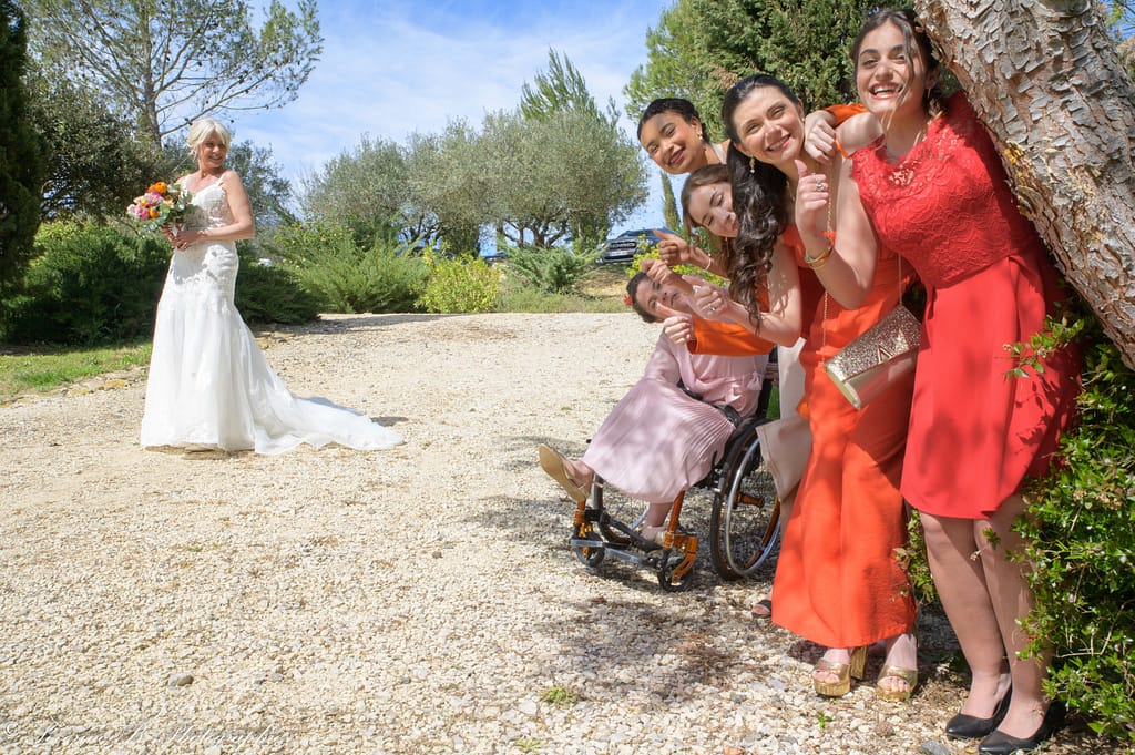 La mariée cherche ses demoiselles d’honneur, toutes cachées derrière un arbre et faisant des signes avec le pouce levé, incluant la demoiselle d’honneur en fauteuil roulant