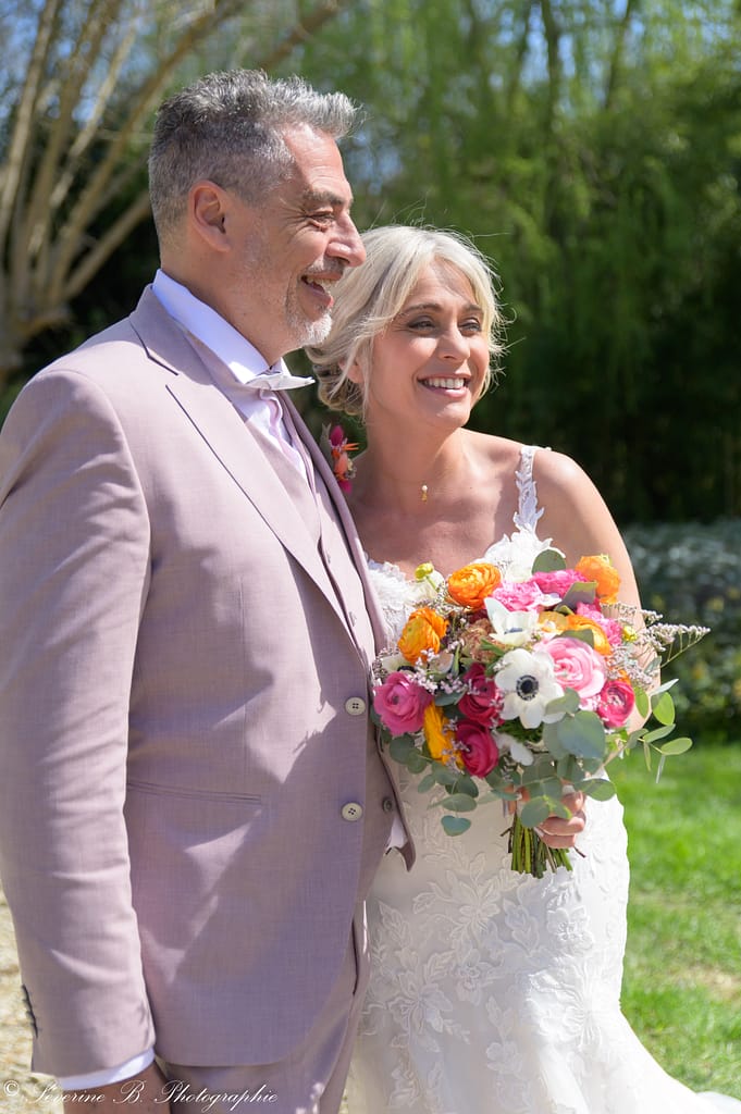 Les mariés, corps l’un contre l’autre, souriants et à l’écoute d’un invité hors‑champ qui les félicite